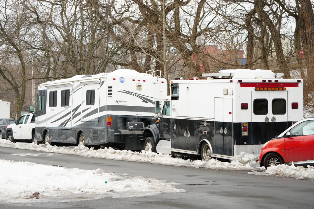 An encampment of RVs and campers along Bronx River Parkway, some lacking license plates or tires, observed operating powered generators, with snow on the ground.