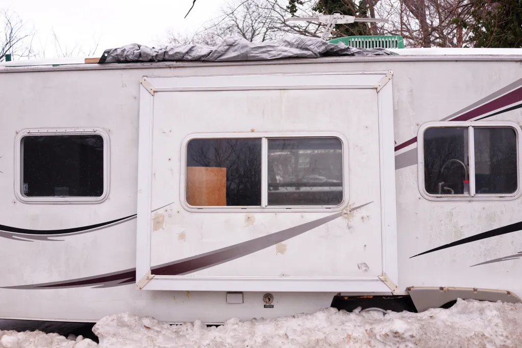 Side view of a white camper or RV with its side extended, a tarp on the roof, and snow piled around its base.