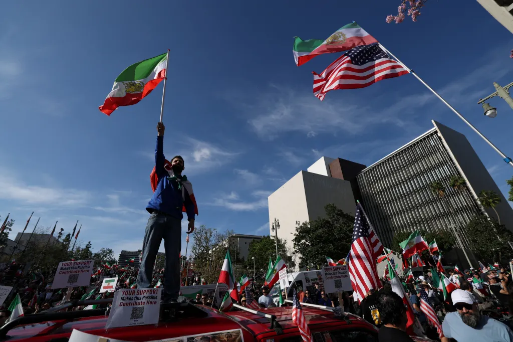 A man stands on a car, holding an Iranian flag, surrounded by a crowd waving Iranian and American flags.
