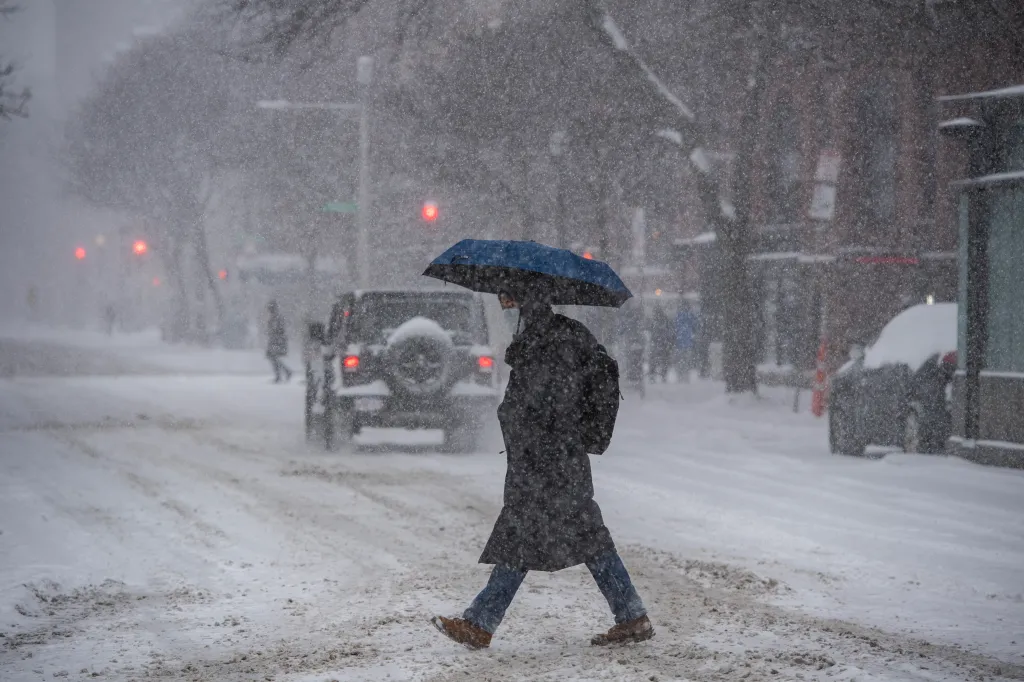 A person in a dark coat and face mask holds a blue umbrella while crossing a snowy street in Boston during a snowstorm.