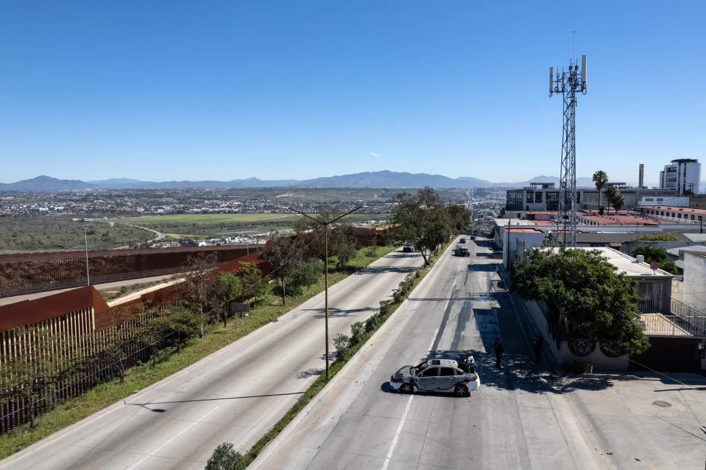 Aerial view of a burnt taxi on a road in Tijuana, Mexico, with the US-Mexico border fence, city, and mountains in the background.