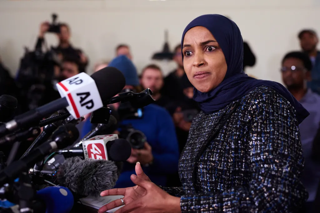 Rep. Ilhan Omar speaking at a news conference, with many microphones in front of her.