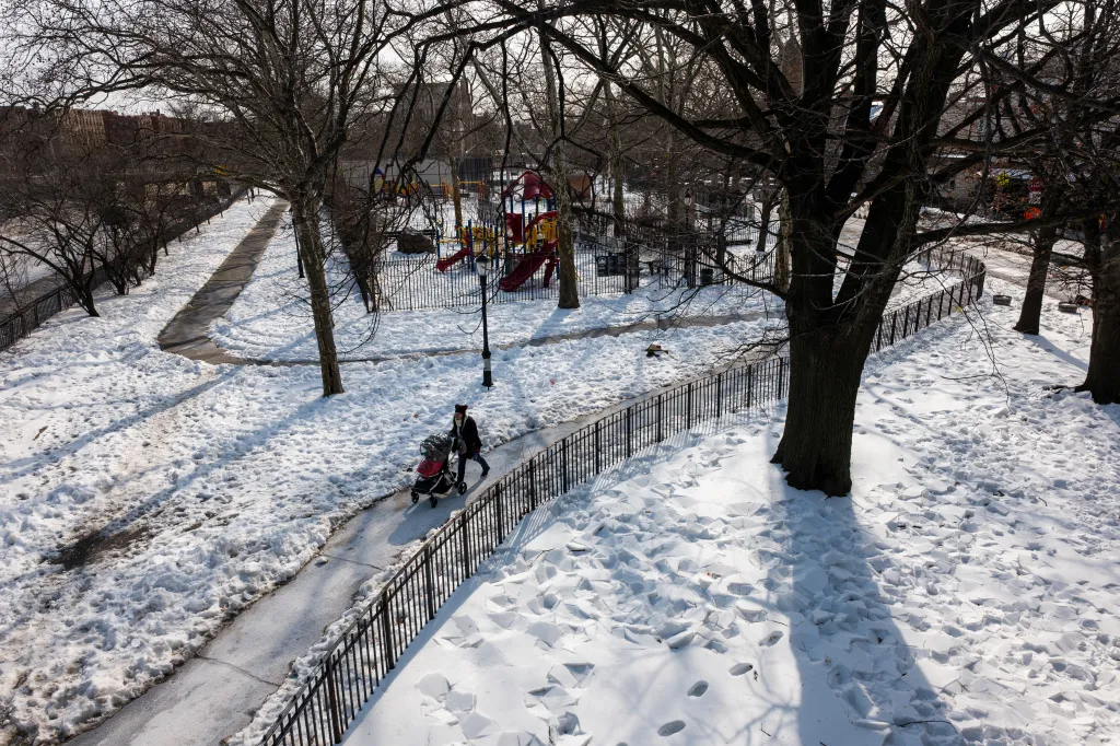A woman bundled in winter clothing pushes a baby stroller on a snow-covered path in a park with a playground.