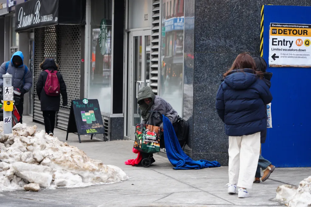 A person experiencing homelessness sits on a New York City sidewalk in extreme cold with melting snow, a Starbucks sign, and a subway detour sign visible.