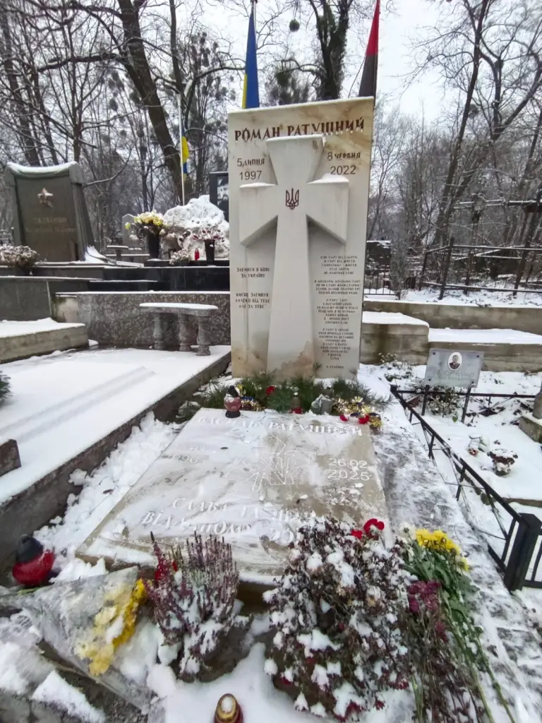 The snow-covered gravesite of Roman Ratushnyi, an independence activist, marked by a cross-shaped headstone and Ukrainian flags.