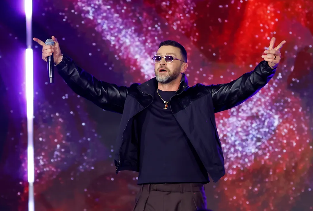 Justin Timberlake performing onstage, wearing sunglasses, a black jacket, and a gold chain, with a red and purple sparkling background.