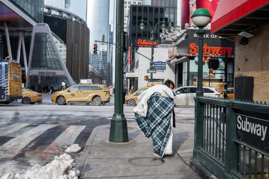 A person wrapped in a large plaid blanket walks past a Subway entrance on a cold day in New York City.
