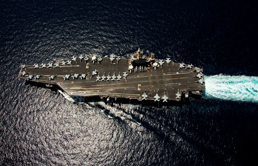 Overhead shot of the USS Abraham Lincoln aircraft carrier in the Arabian Sea with numerous fighter jets on its deck, leaving a white wake behind it.