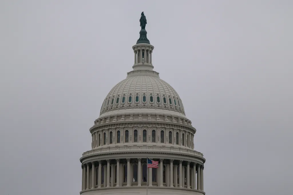 The dome of the U.S. Capitol building with an American flag flying below it, under a gray sky.