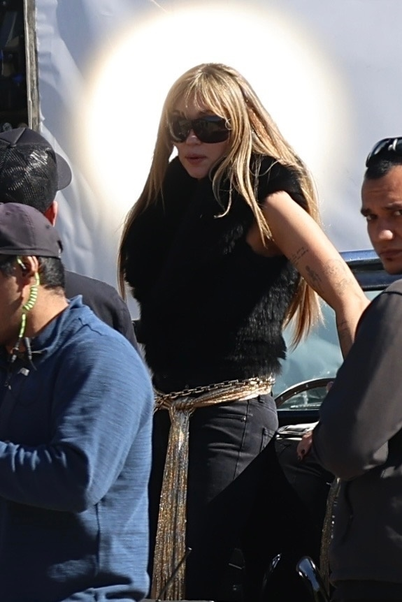 Miley Cyrus with blonde hair and sunglasses, wearing a black furry top, black pants, and a gold chain belt, filming the Hannah Montana 20th Anniversary Special in Malibu.