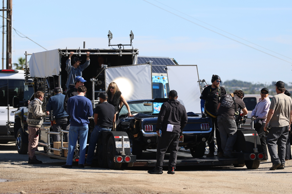 Miley Cyrus as Hannah Montana with blonde hair and sunglasses filming scenes for the Hannah Montana 20th Anniversary Special in a Ford Mustang.