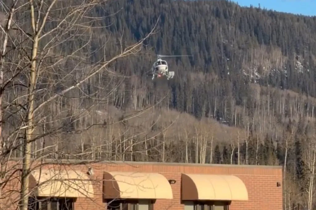 A helicopter flying over a brick building with mountains and bare trees in the background.
