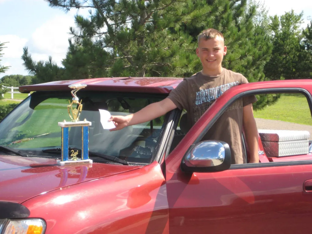 A young man smiles while holding paper and standing next to a red truck with a