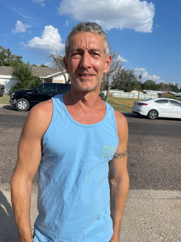 A man with gray hair and a blue tank top stands on a paved road with cars and houses in the background.