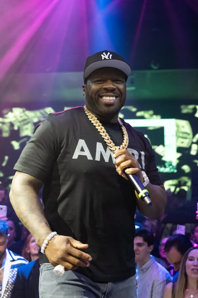 Rapper 50 Cent performing on stage in a black shirt, a black New York Yankees baseball cap, and a large gold chain, holding a microphone.
