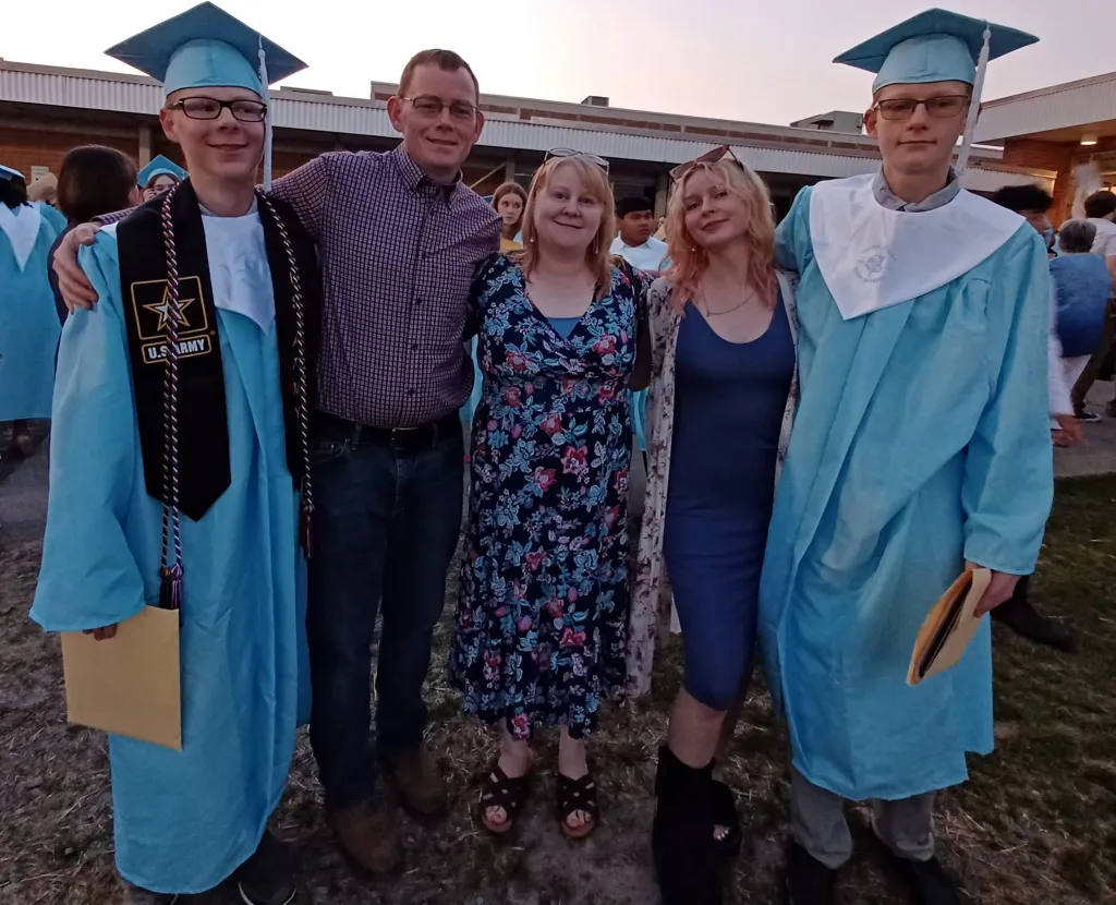 Two graduates in light blue gowns and caps stand with three family members.