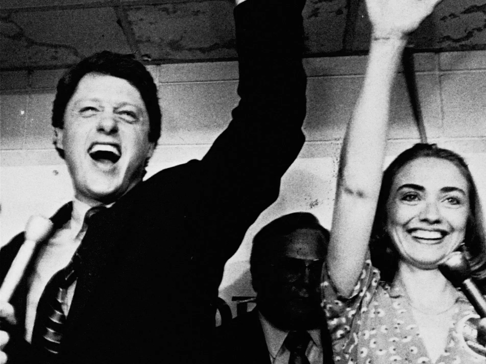 A black and white photo of Bill Clinton and Hillary Clinton smiling with their arms raised.