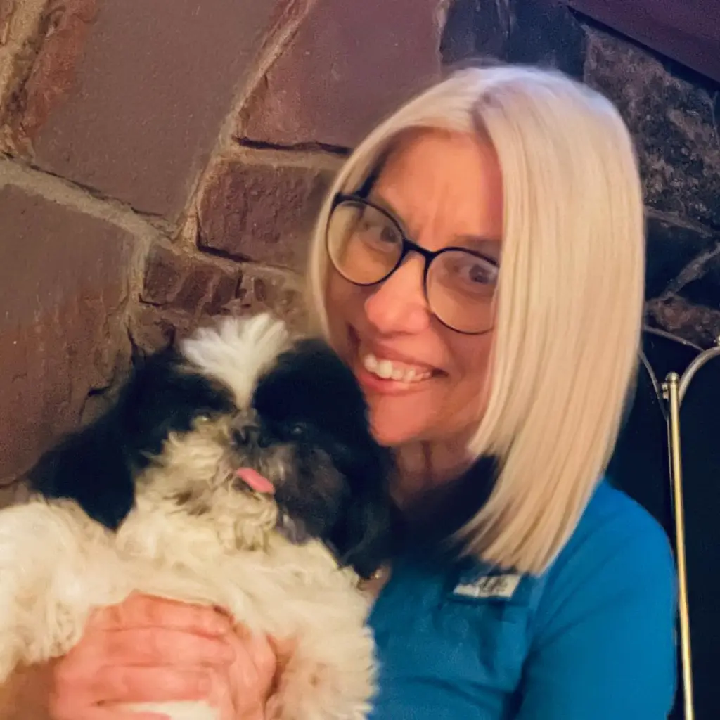 A smiling woman with blonde hair and glasses holding a fluffy black and white dog.