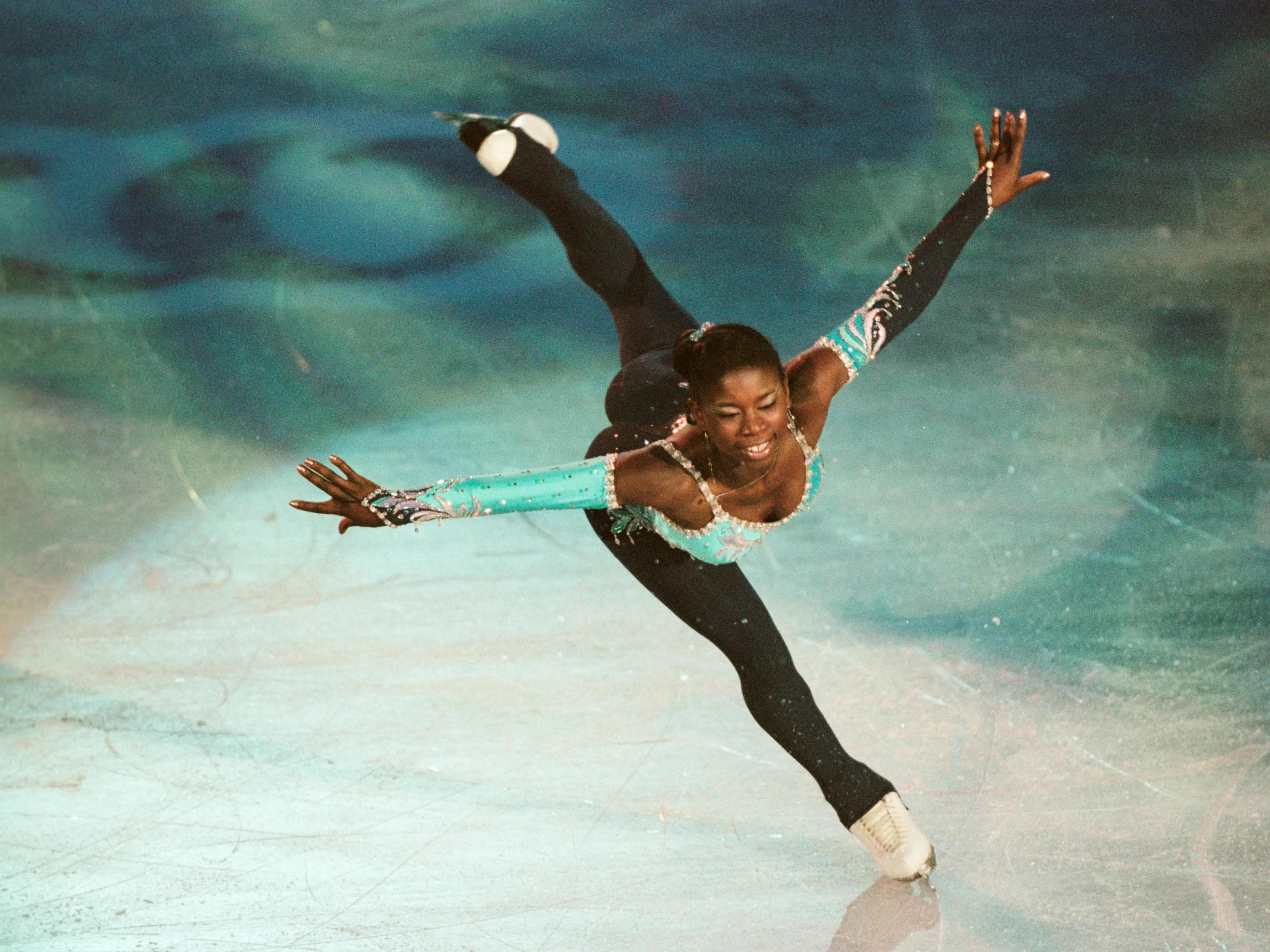 Surya Bonaly skating with her arms extended and one leg raised behind her. She wore a full-body black and turquoise costume.