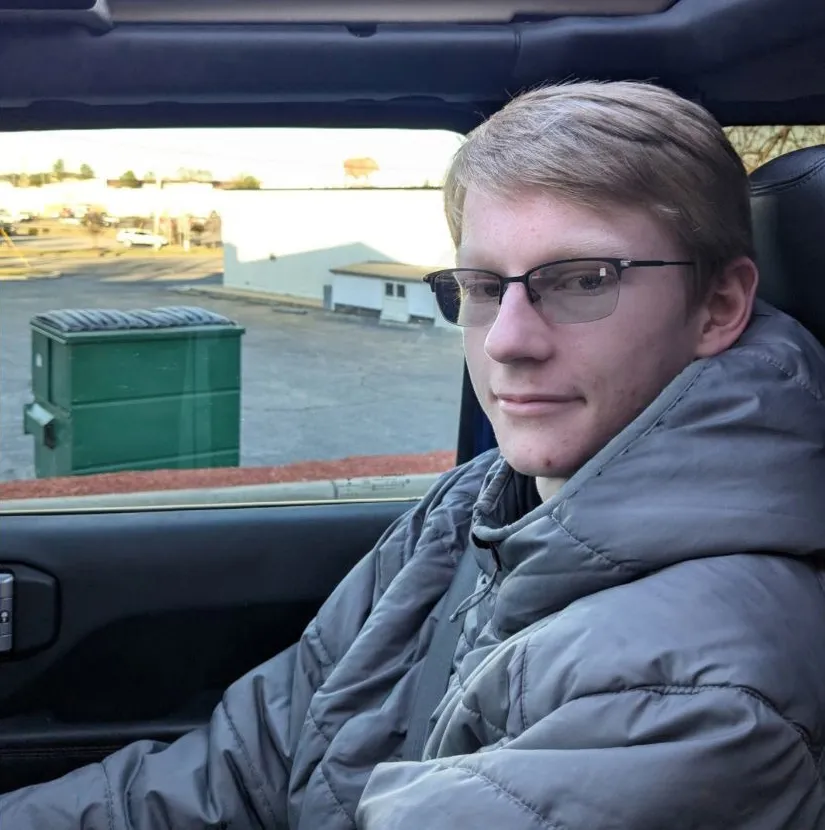 Young man in a gray puffer coat and glasses looking out a car window.