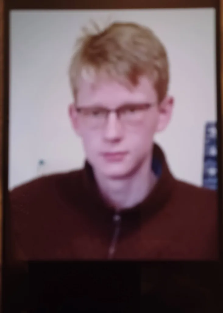 A blurry portrait of a young man with light brown hair and glasses, wearing a dark brown jacket.