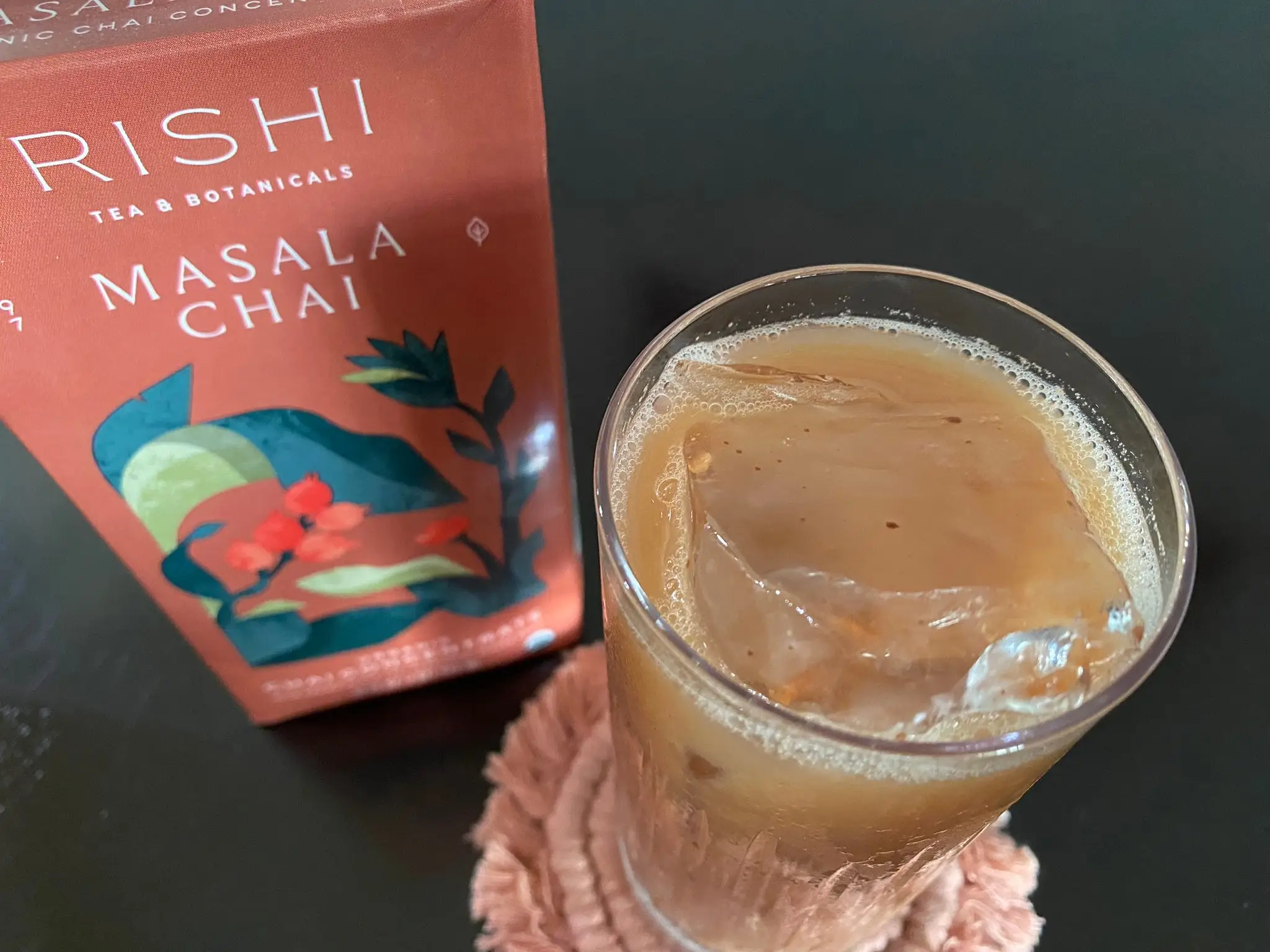 Carton of Rishi cha concentrate next to glass of iced chai latte and spice-flecks floating in drink.