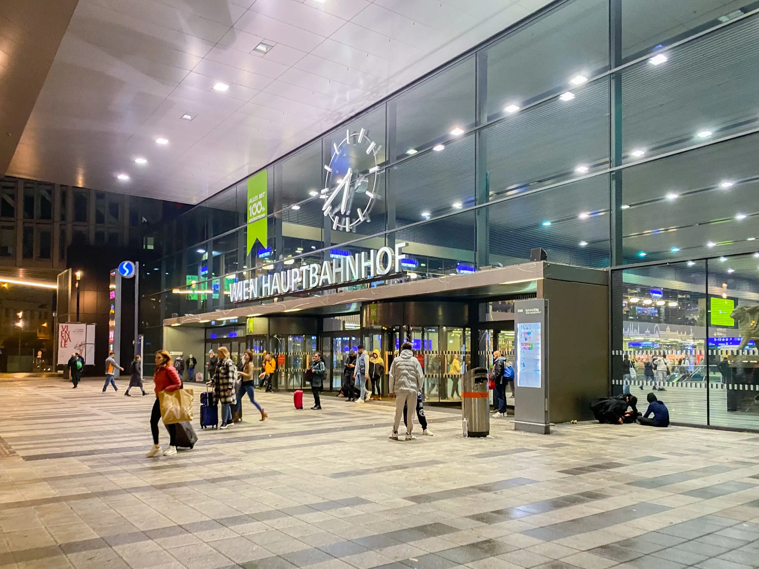People with suitcases walk outside of a train station at night.