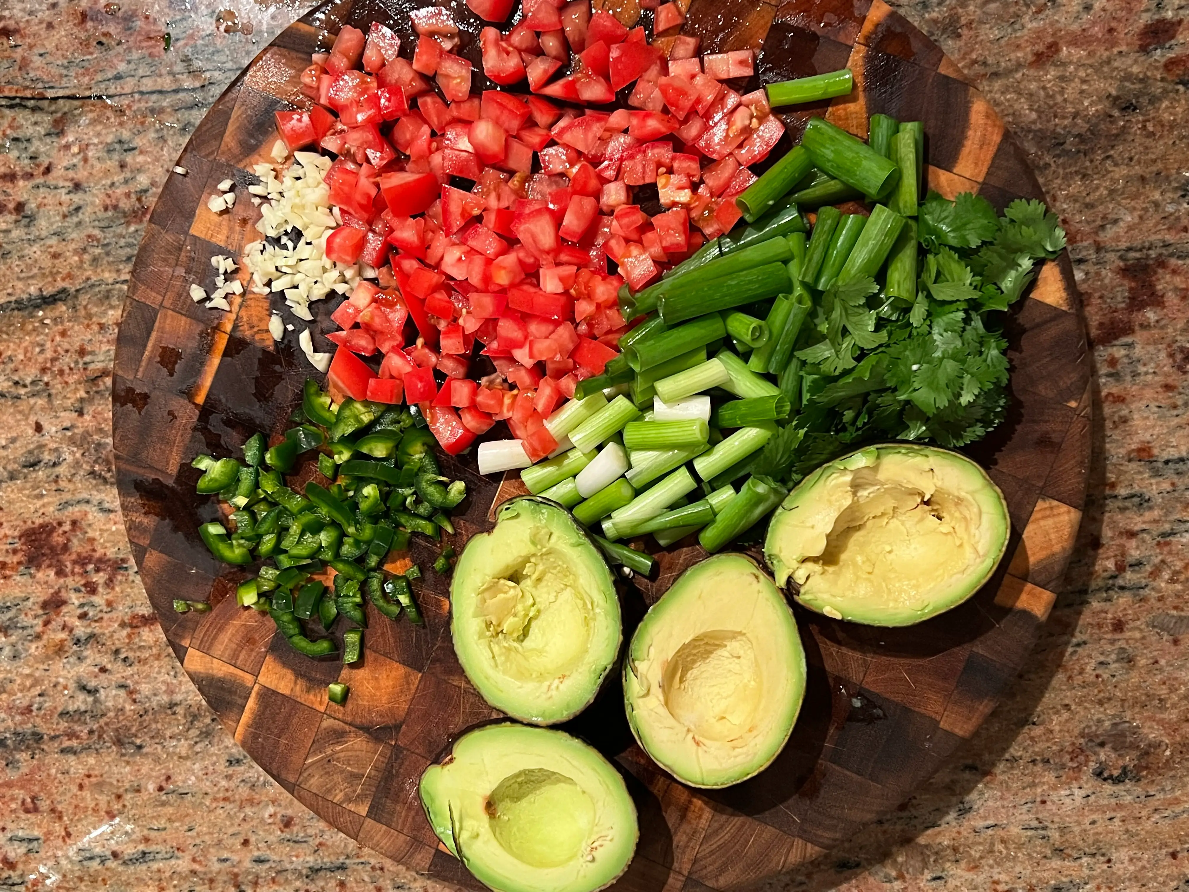 A cutting board with diced garlic, peppers, cilantro, scallions, tomatoes, and halved avocados