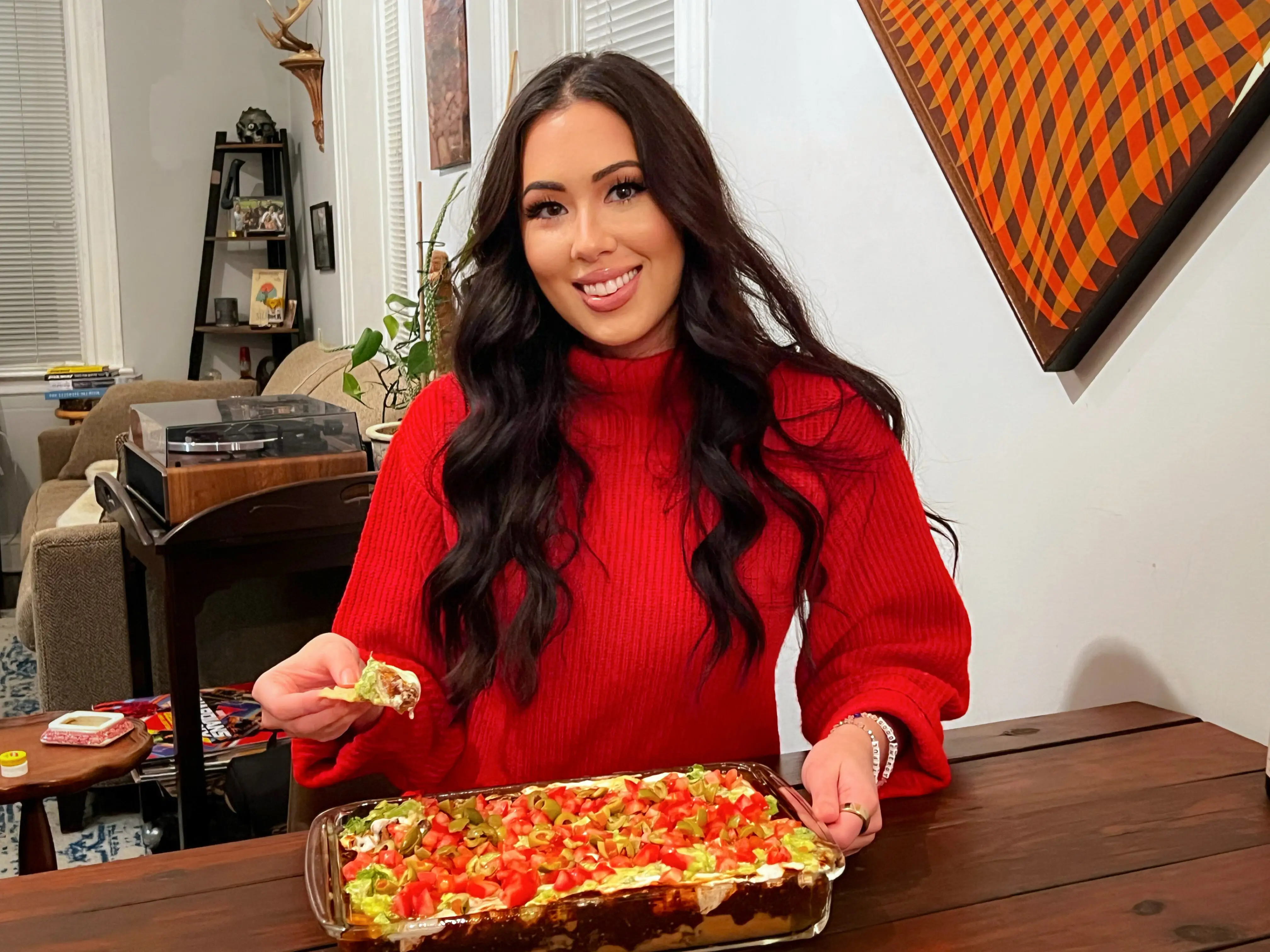 The writer, wearing a red shirt, smiles and holds a tortilla chip with 7-layer dip on it. A casserole dip garnished with tomatoes, olives, and guacamole sits in front of her