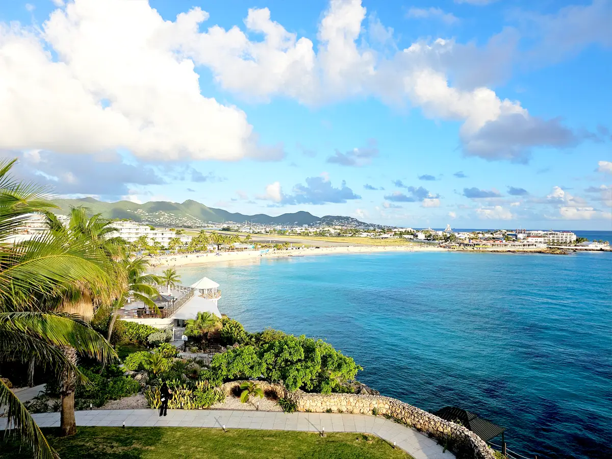 A beach with deep-blue water next to a pathway surrounded by palm trees and greenery and houses and mountains in the background.