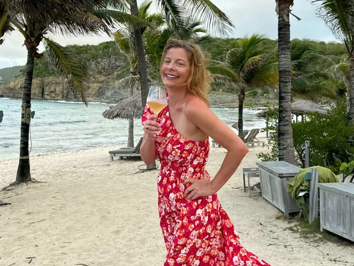 The writer wears a long floral dress and holds a cocktail in front of palm trees and a beach.
