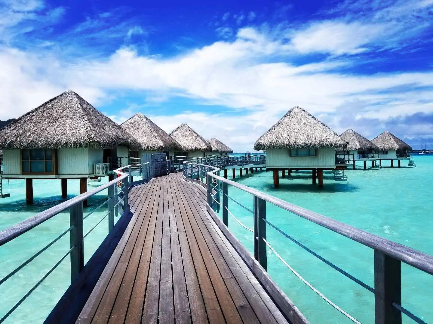 A dock leading to at least seven bungalows with straw rooves sitting above a bright aqua lagoon in Bora Bora.