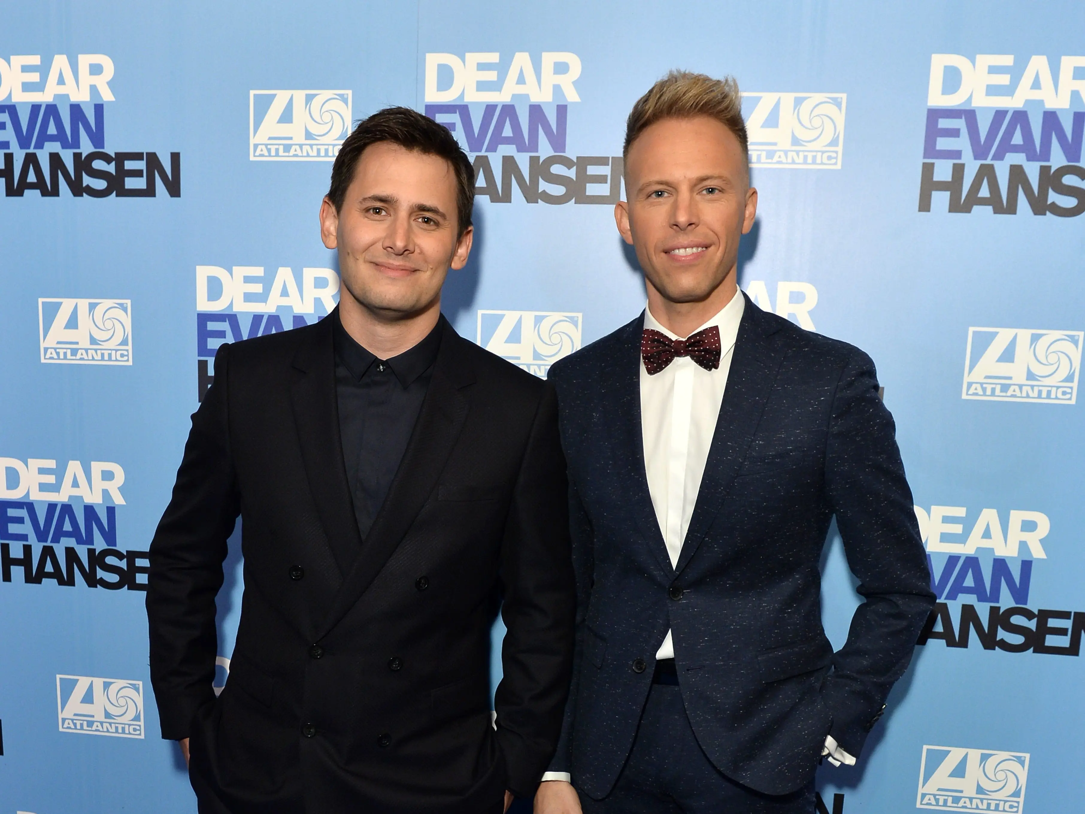 benj pasek, a man with short hair wearin g a black suit and black shirt, next to justin paul, wearing a navy suit, white shirt, and red bowtie, on a blue carpet at a dear evan hansen premiere