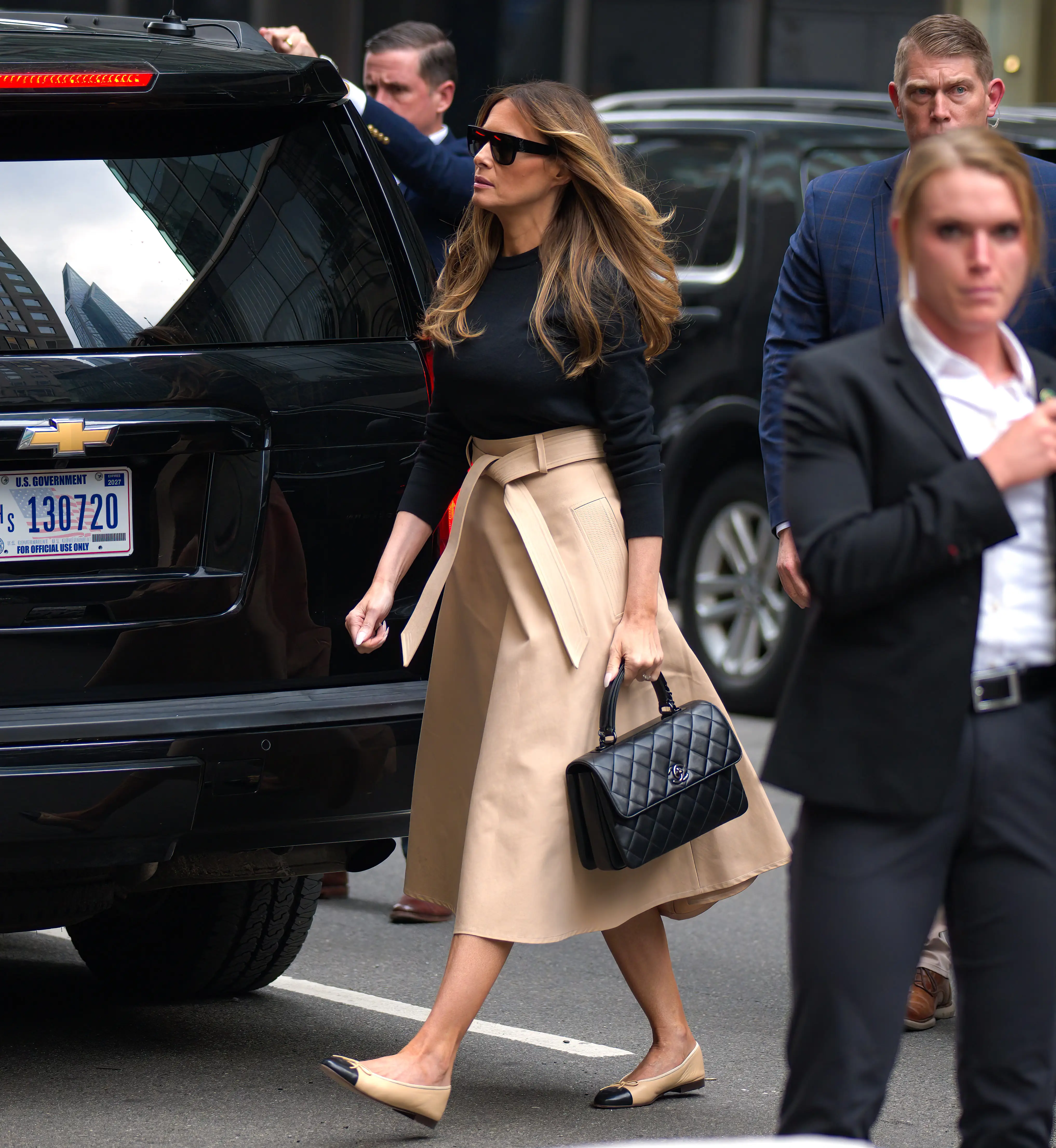 Melania Trump walks down the street in a black top and tan skirt.
