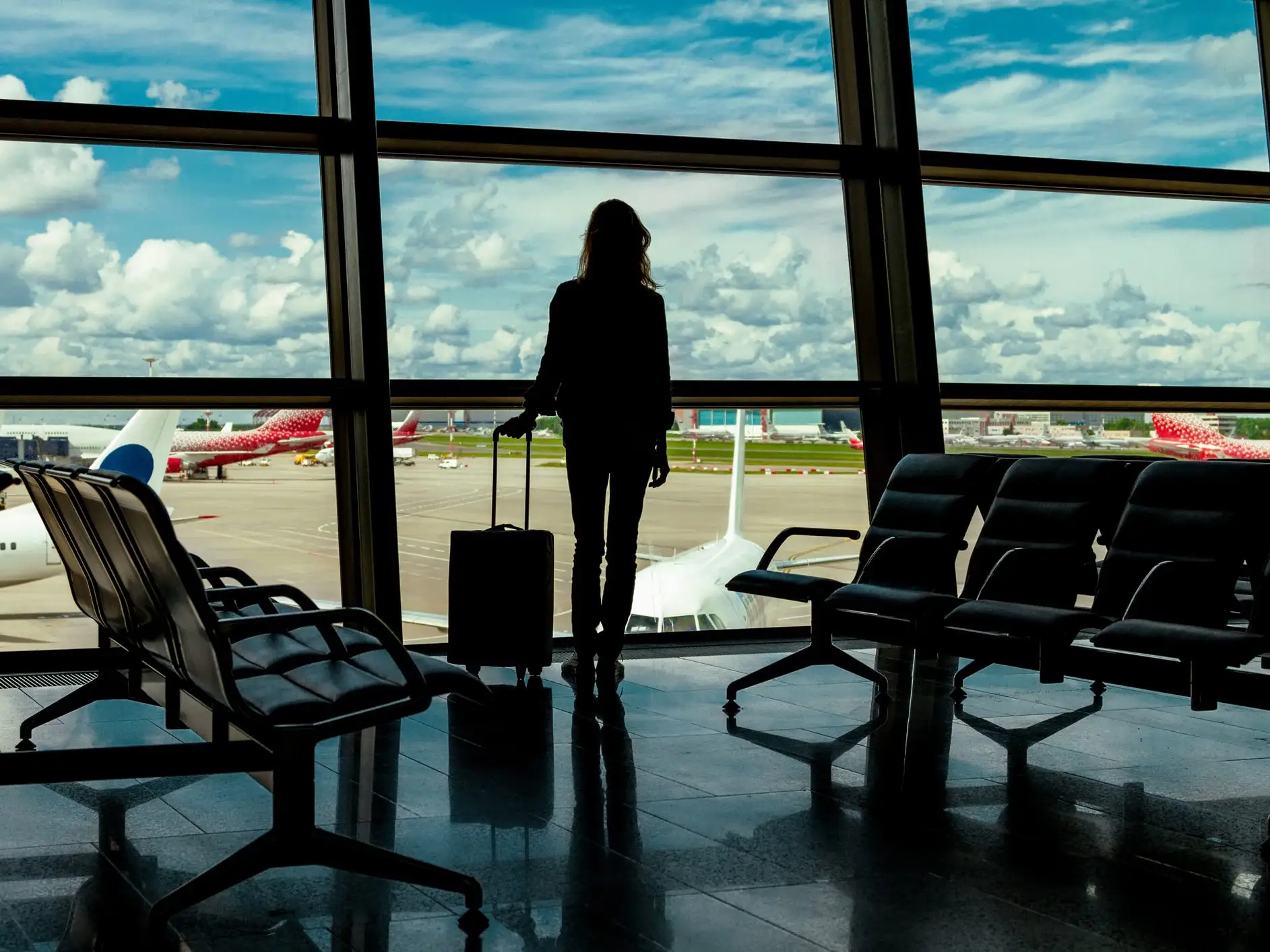 Silhouette of a woman with a carry-on bag surrounded by seats looking out onto a runway at an airport