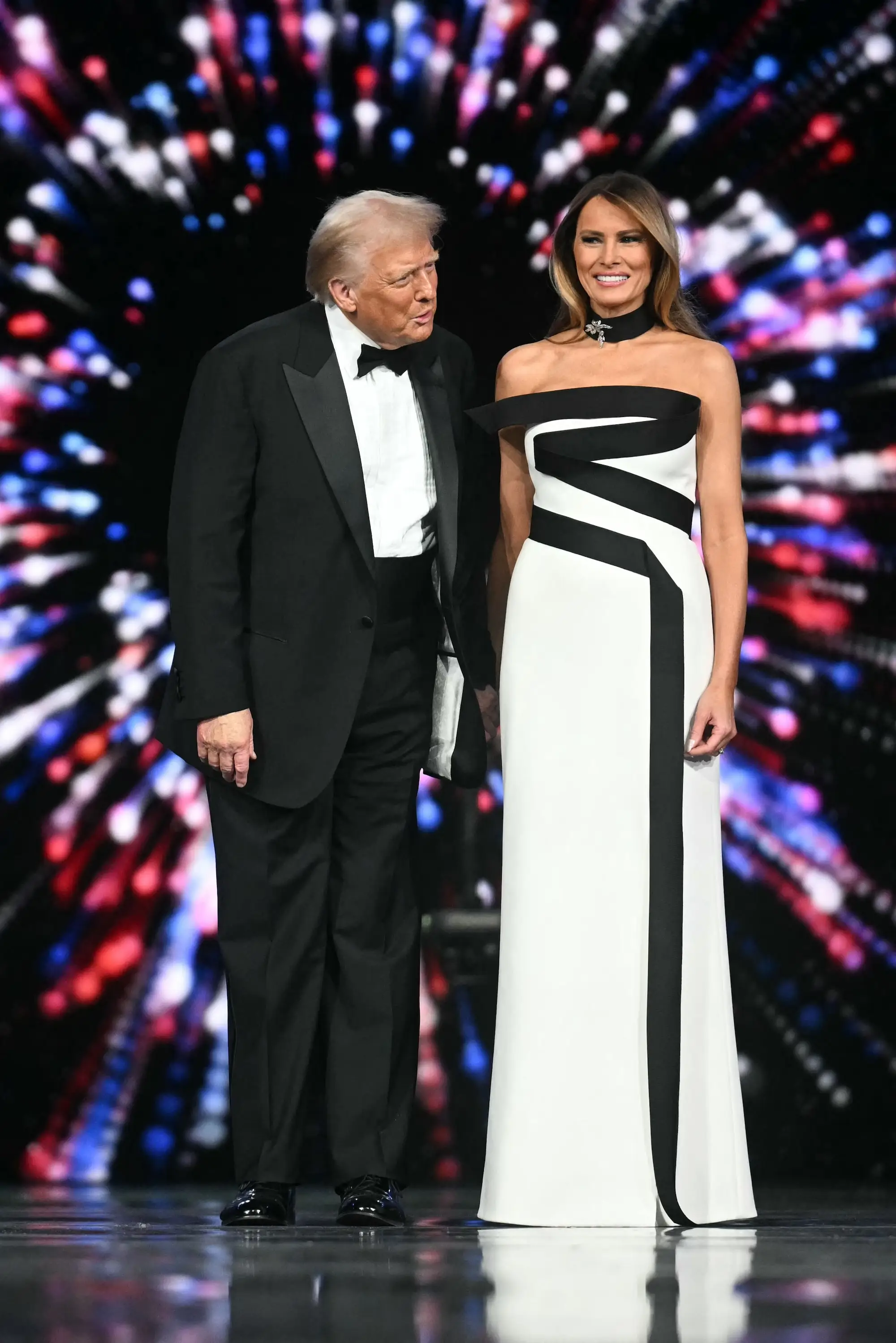 Donald Trump and Melania Trump onstage at the 2025 Inaugural Ball, with a screen showing red, white, and blue lights behind them. He's wearing a black tux, and she's wearing a white strapless gown with a black strip of fabric zig-zagging across the bodice and straight down the skirt,