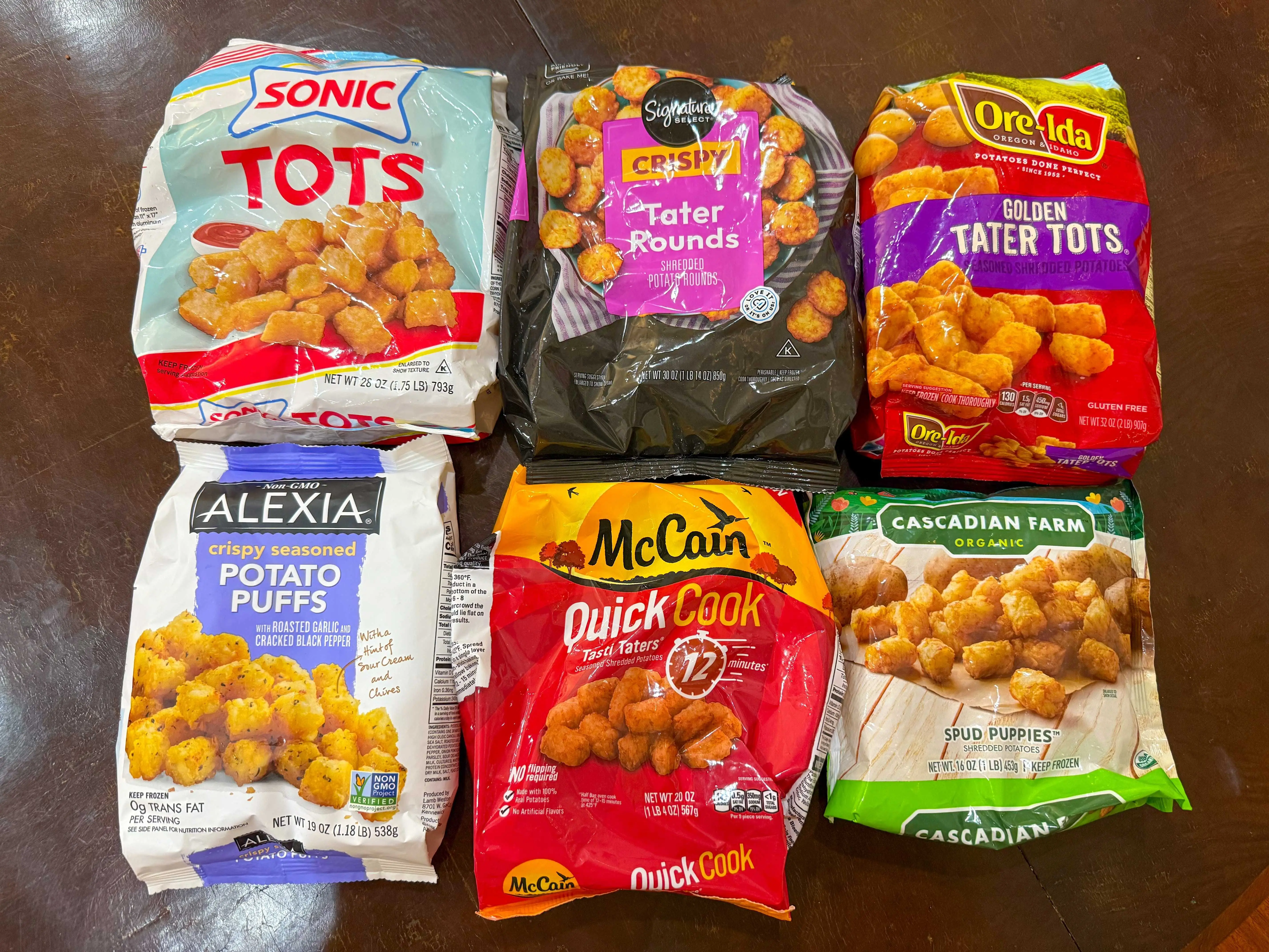 Six bags of tater tots from brands including Sonic, Signature Select, Ore-Ida, Alexia, McCain, and Cascadian Farm on a brown table