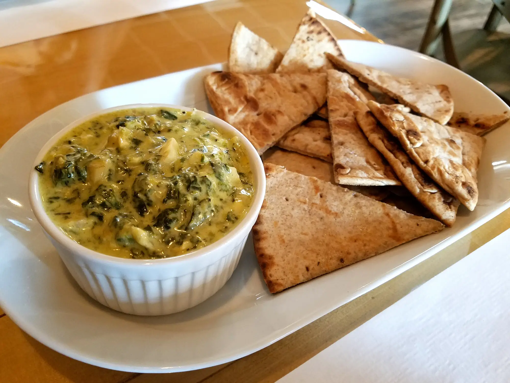 A white oval shaped dish holding a white ramekin filled with spinach-artichoke dip and and pita chips