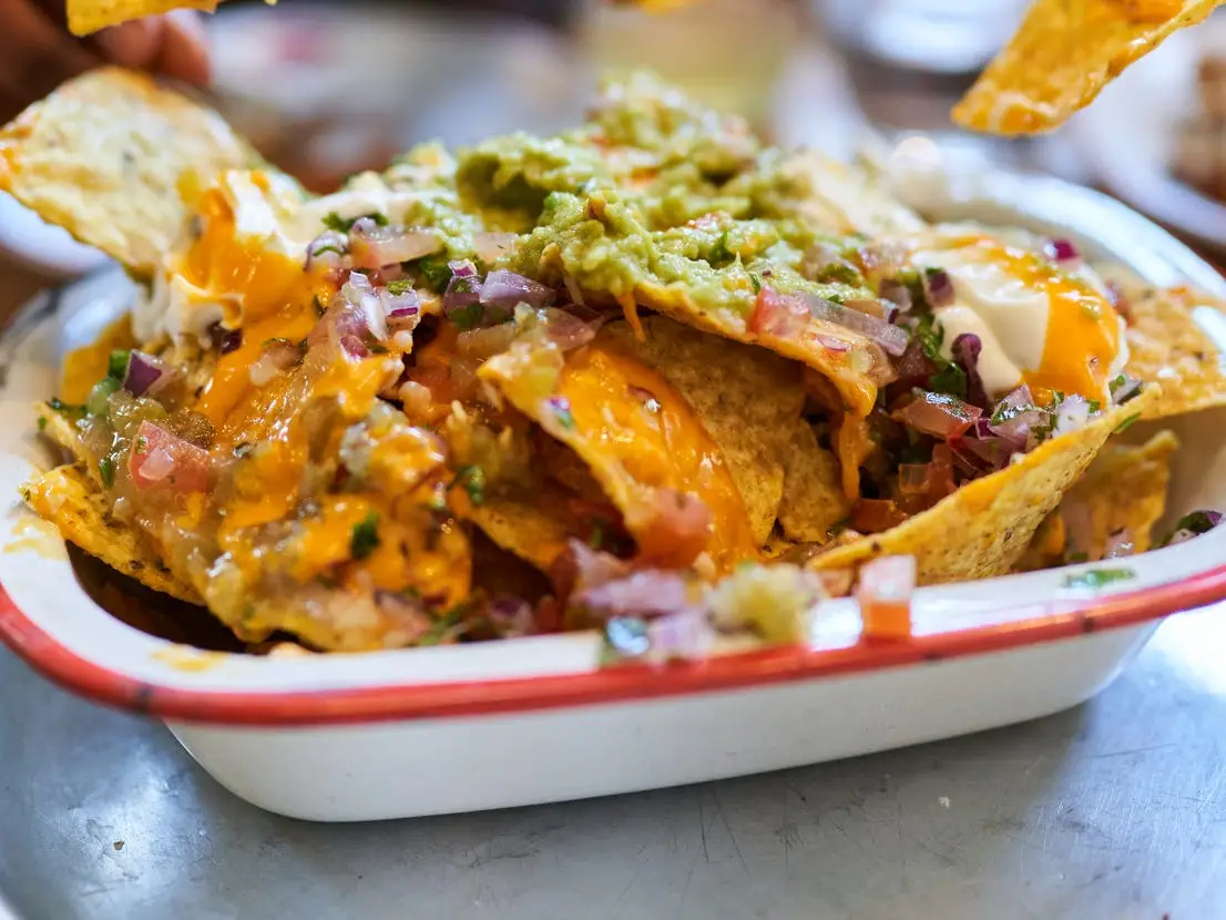 A white and red tray serving dish filled with nachos covered in cheese, onions, tomatoes, and guacamole at a table
