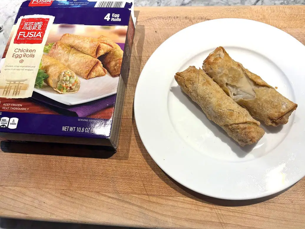 Two egg rolls on a white plate next to the box the product came in.