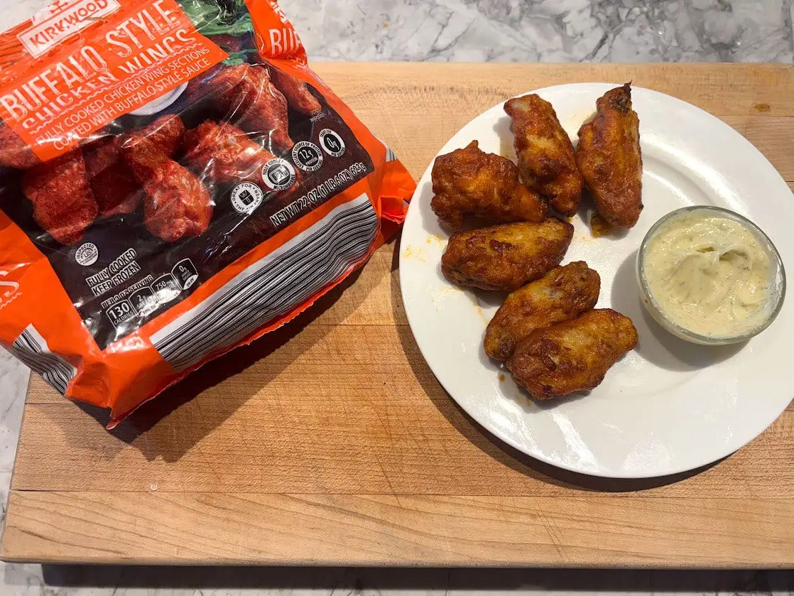 Six Buffalo wings and a small bowl of sauce on a white plate next to the box the product came in.