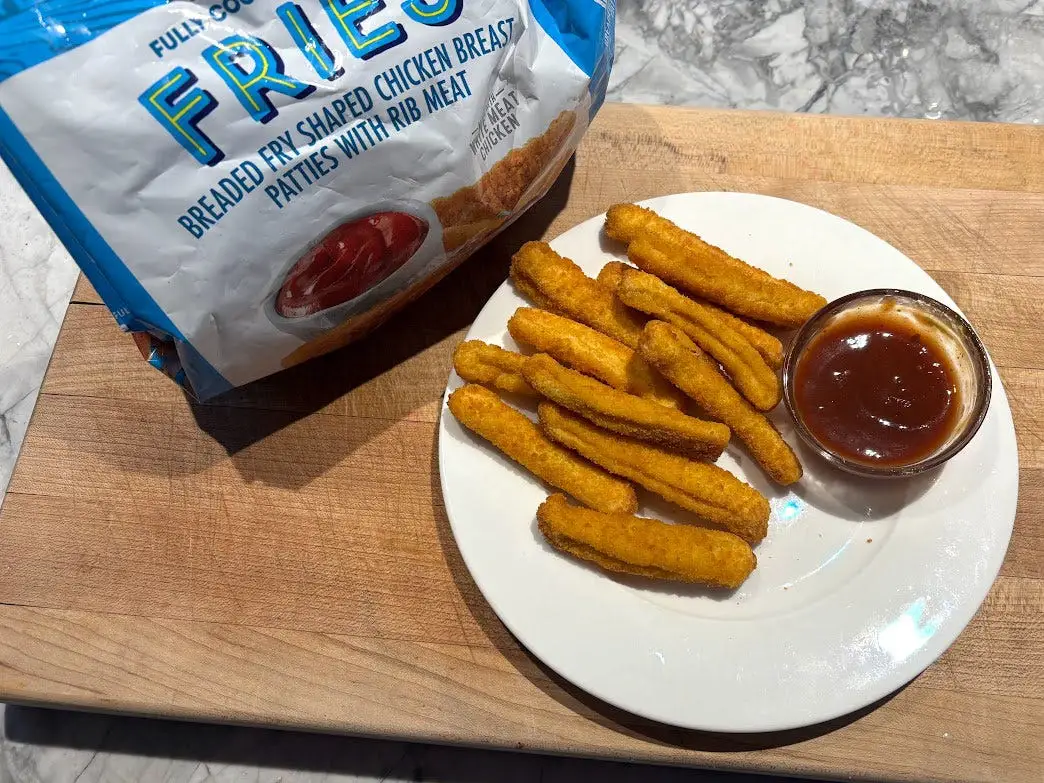 Chicken fries and a small bowl of sauce on a white plate next to the packaging the product came in.
