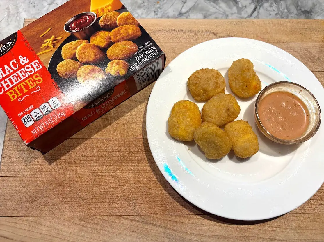 Mac and cheese bites and a small bowl of sauce on a white plate next to the box the product came in.