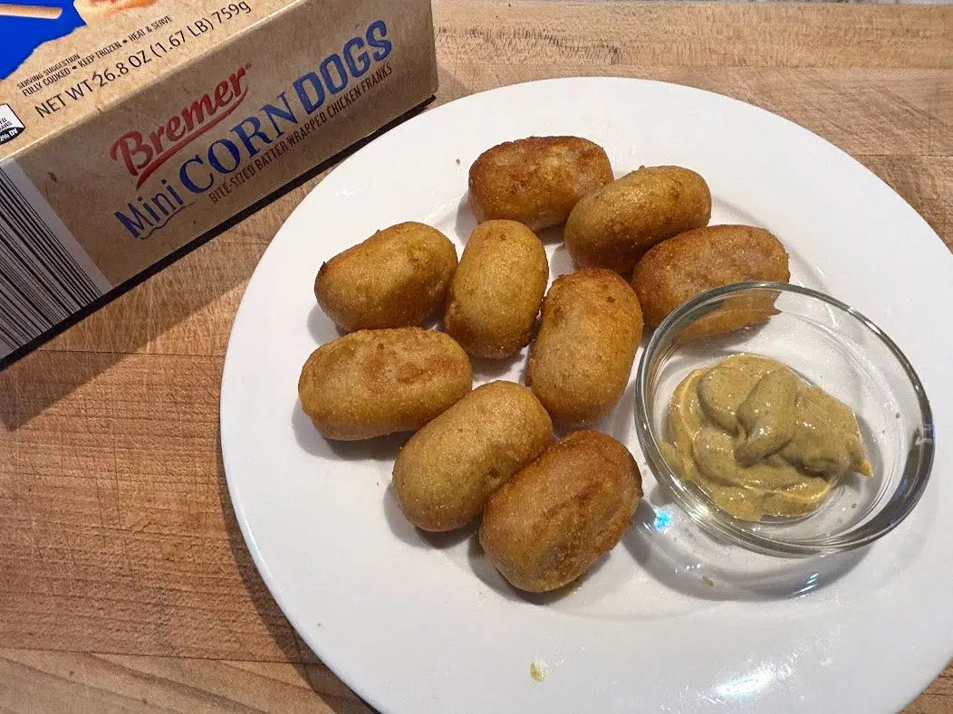 Mini corn dogs and a small bowl of mustard on a white plate next to the packaging the product came in.