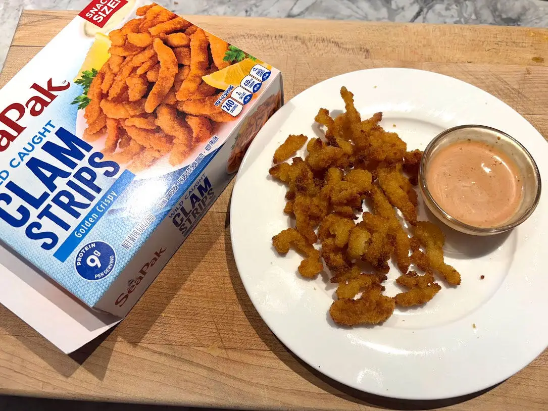 Clam strips and a small bowl of sauce on a white plate next to the packaging the product came in.