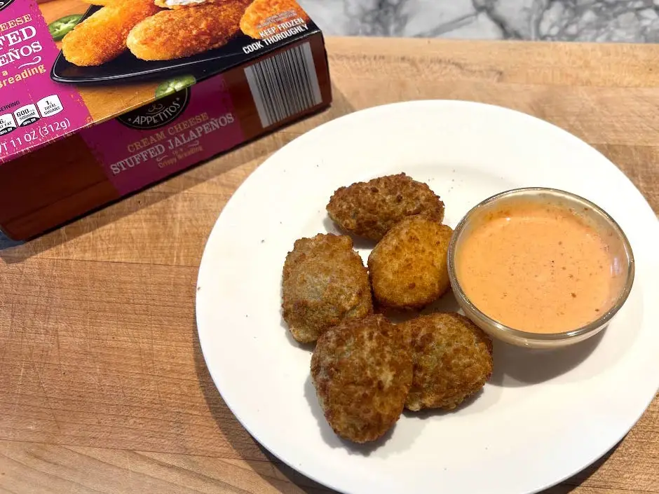 Five jalapeño poppers and a small bowl of sauce on a white plate next to the packaging the product came in.