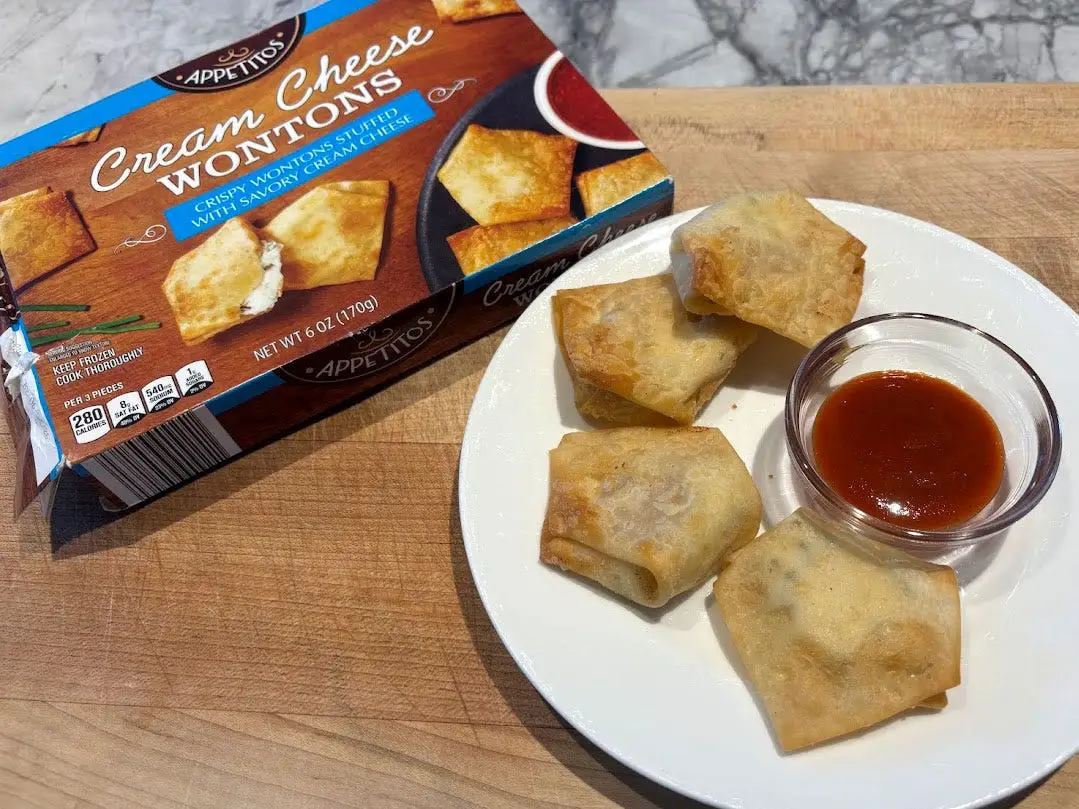 Four wontons and a small bowl of sauce on a white plate next to the packaging the product came in.