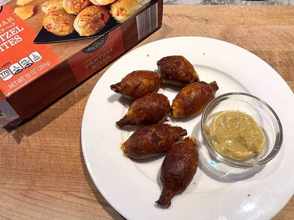 Mini pretzel bites and a small bowl of mustard on a white plate next to the packaging the product came in.