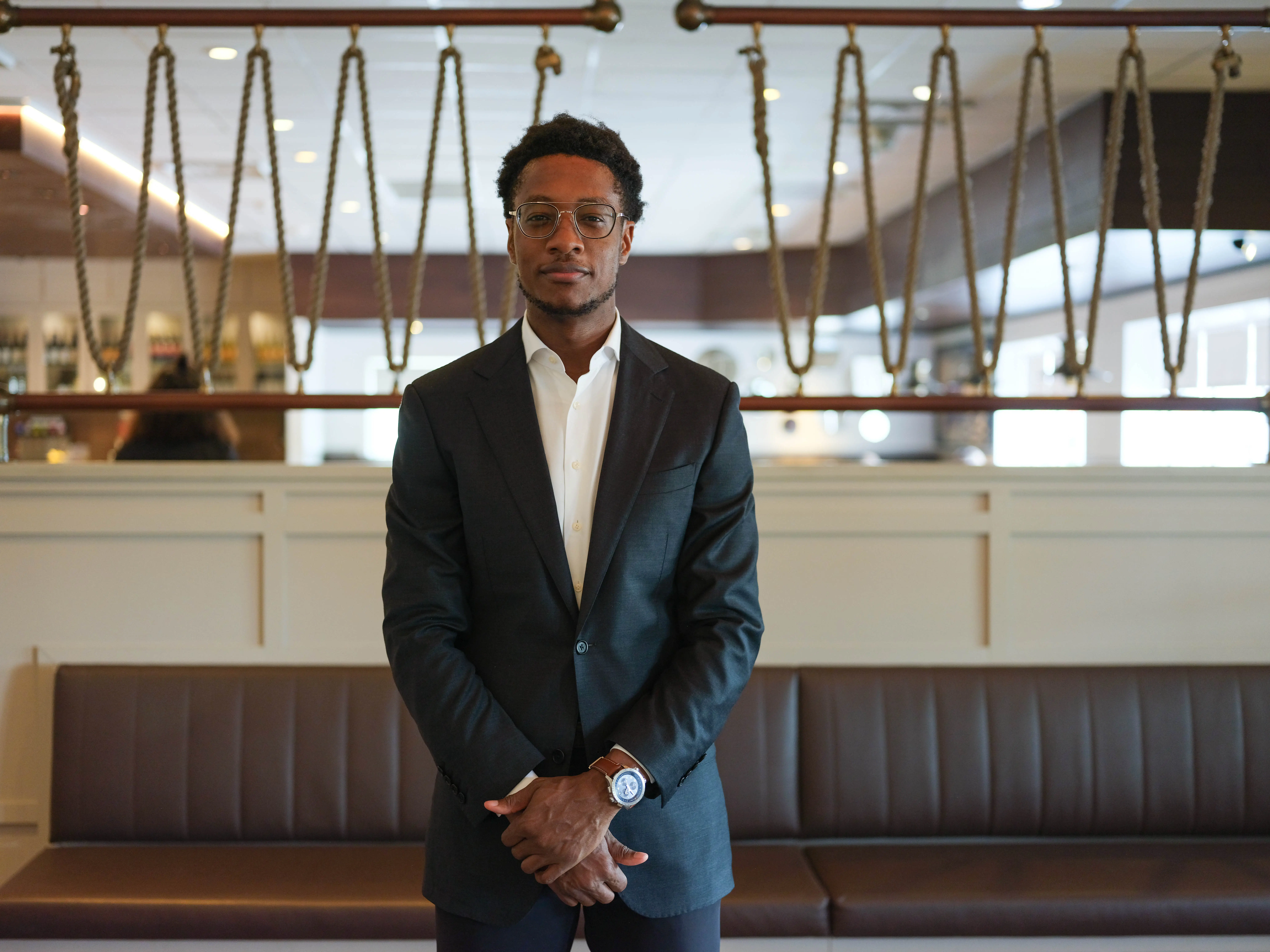 Damola Adamolekun standing in suit with hands folded in front of a restaurant bench.
