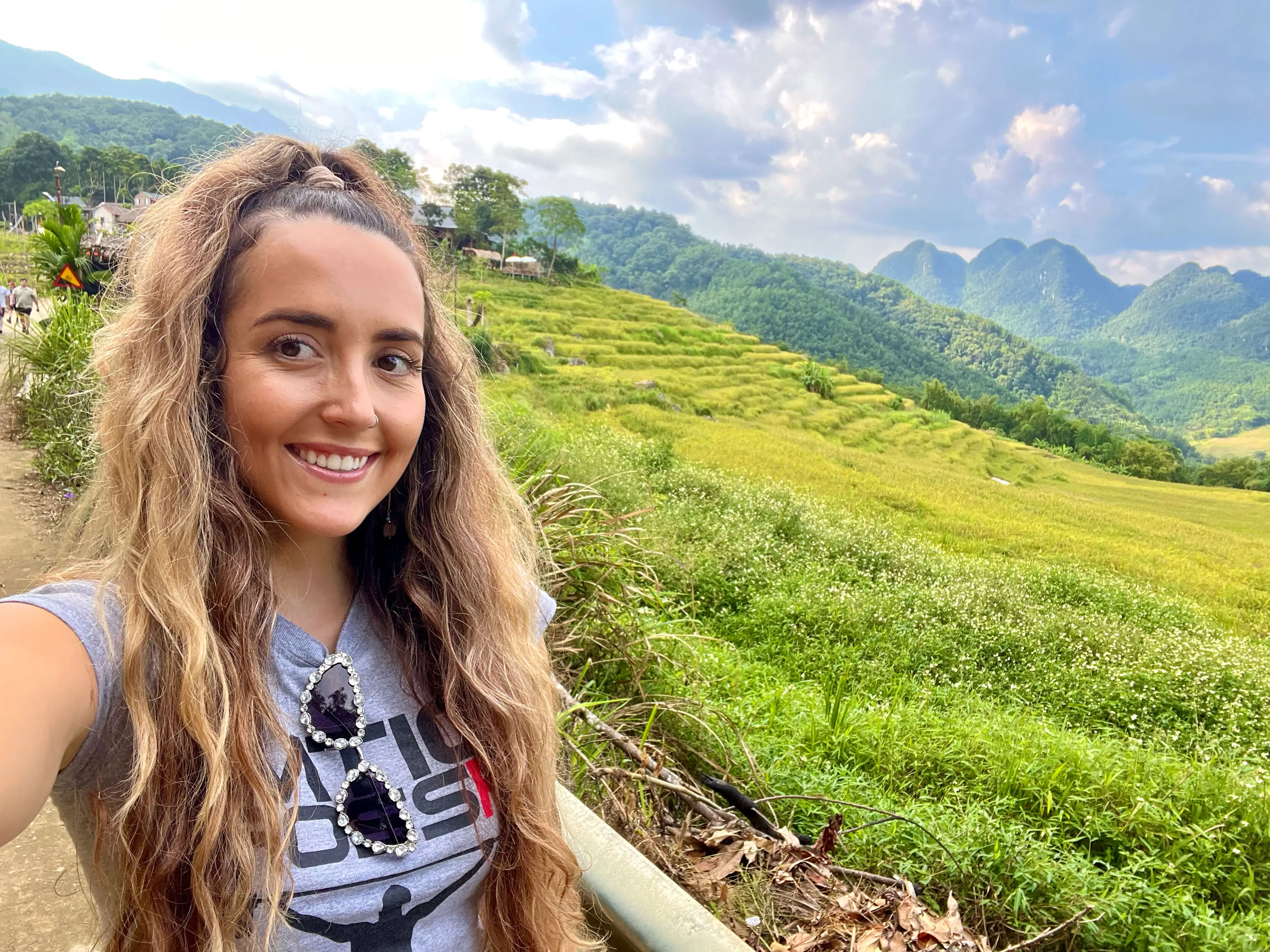 A woman takes a selfie in front of grassy fields and mountains on a cloudy day.